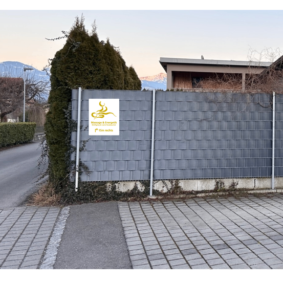 Ein grauer Holzzaun mit Logo und dem Text Massage & Energietechnik steht vor einem Gebäude mit Bergen im Hintergrund.