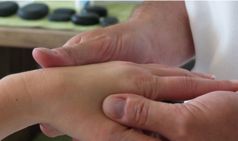 Eine erwachsene Hand hält die Hand eines Kindes, möglicherweise für eine Massage. Im Hintergrund sieht man einen Holztisch mit mehreren schwarzen Steinen.