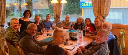 Eine Gruppe von Menschen, sowohl Männer als auch Frauen, sitzen um einen Tisch in einem Restaurant und lächeln für ein Foto. Der Tisch ist mit Getränken und Essen gedeckt, und der Raum hat ein großes Fenster.