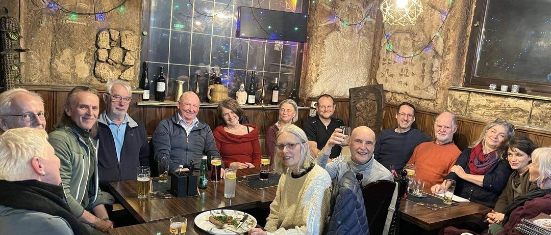 Eine Gruppe von Menschen in einer Kneipe lächelt und posiert für ein Foto. Sie sitzen um Holztische herum, auf denen verschiedene Getränke und Teller stehen.