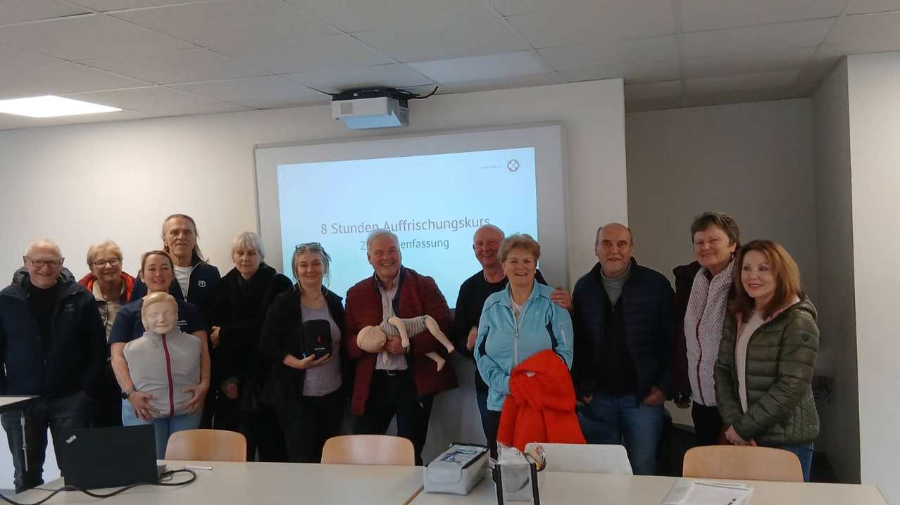 A group of adults stands in a classroom, smiling for a photo. A projector screen displays an 8-hour resuscitation course. A man holds a dummy, and a woman holds a black case.