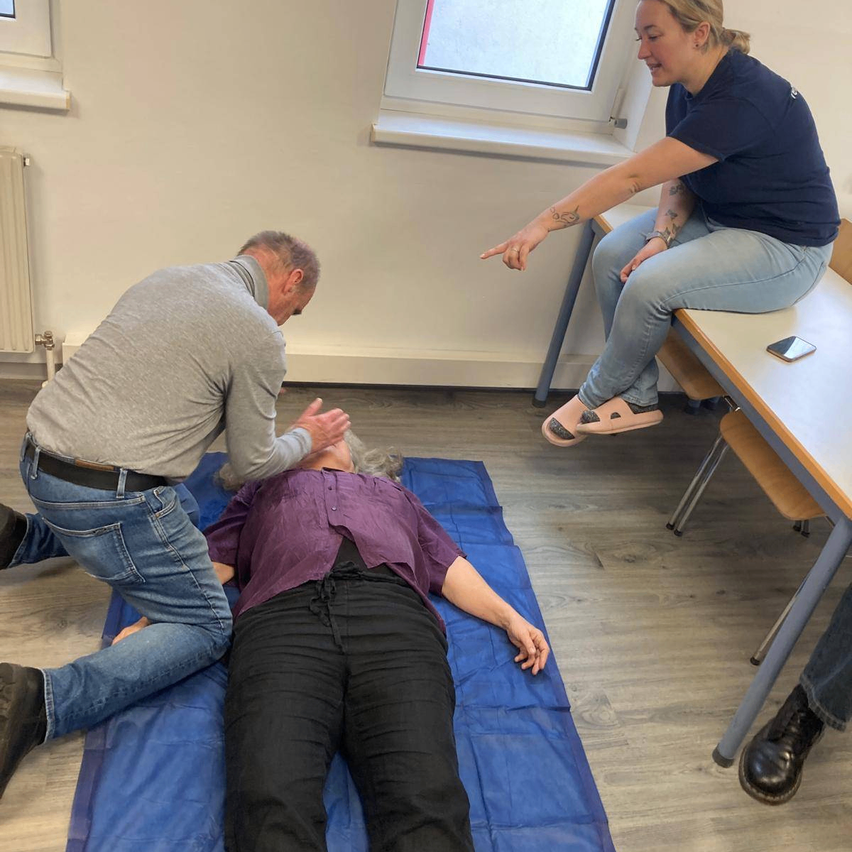 A man kneels over a woman lying on a blue mat while another woman sits at a table and points. The room has wooden floors and white walls.