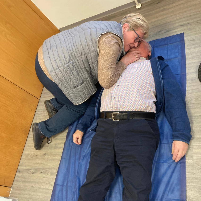 An elderly man lies on a blue mat, being comforted by a woman wearing glasses.