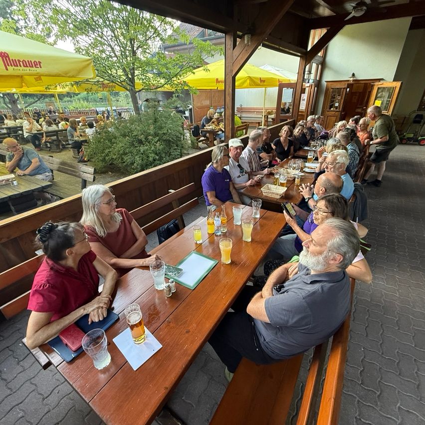 Eine Gruppe von Menschen sitzt an einem Tisch in einem Restaurant im Freien, trinkt Bier und führt ein Gespräch. Das Restaurant hat Holztische und Stühle, und im Hintergrund befinden sich Schirme und ein Baum.