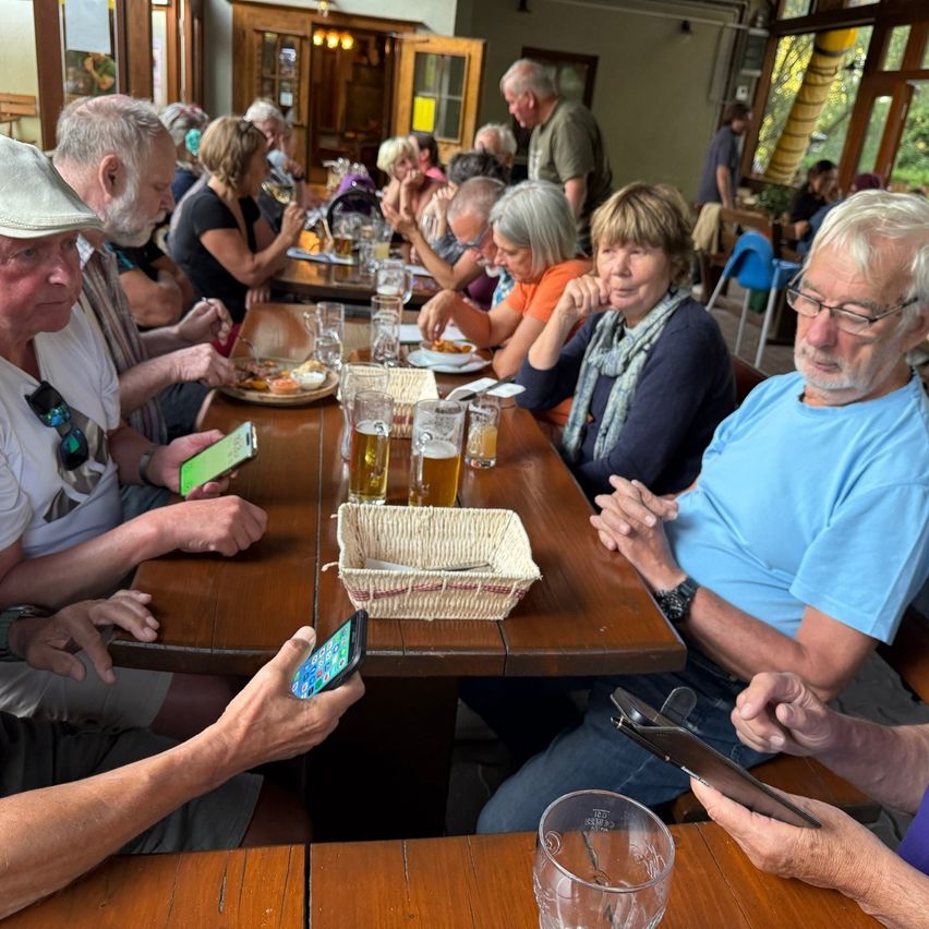 Eine Gruppe älterer Menschen sitzt an einem Tisch in einem Restaurant, einige essen, während andere ihre Handys benutzen. Auf dem Tisch stehen Gläser Bier und Teller mit Essen.