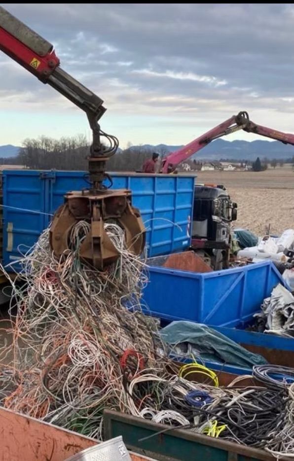 Ein Kran hebt einen großen Haufen verhedderter Drähte an, die in blauen Müllcontainern platziert sind, unter einem bewölkten Himmel.