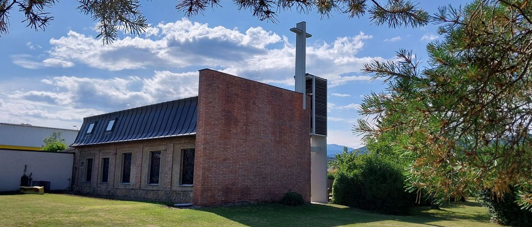 Eine Kirche mit Backsteinfassade, einem Kreuz oben und Fenstern. Die Kirche ist von Büschen und Gras umgeben.