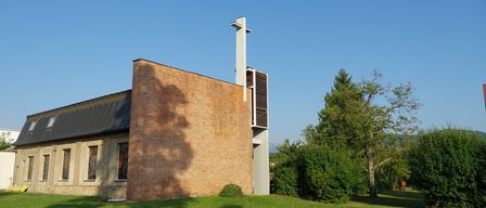 Ein modernes Backsteingebäude mit einem hohen weißen Turm und einem Kreuz darauf, umgeben von Grün unter einem klaren blauen Himmel.