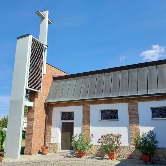 Eine moderne Kirche mit Ziegelwänden und einem Metalldach, ausgestattet mit einem hohen Glockenturm. Der Eingang wird von Topfpflanzen und Glasfenstern flankiert. Der Himmel ist klar mit einigen Wolken.