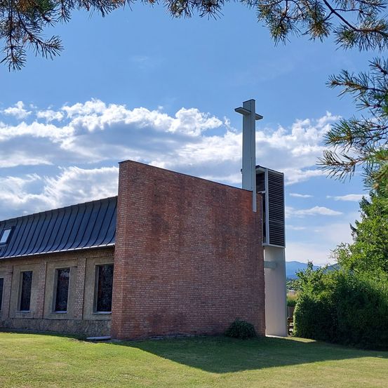 Eine Kirche mit einer Ziegelmauer, einem grauen Dach und einem Kreuz oben. Sie hat mehrere Fenster und ist von Grün umgeben.
