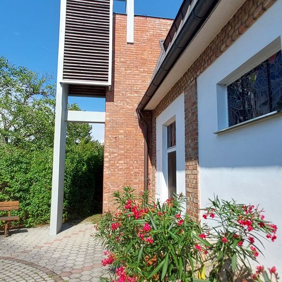 Ein Backsteingebäude mit einem weißen Pfeiler und einem Glasfenster mit Design. Es hat eine Glastür mit weißem Rahmen. Vor dem Gebäude befinden sich Blumen und Pflanzen. Links steht eine Bank.