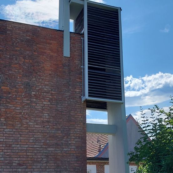 Ein hoher, schlanker Backsteinbau mit einer weißen, gitterartigen Struktur an seiner Seite, unter einem teilweise bewölkten blauen Himmel.