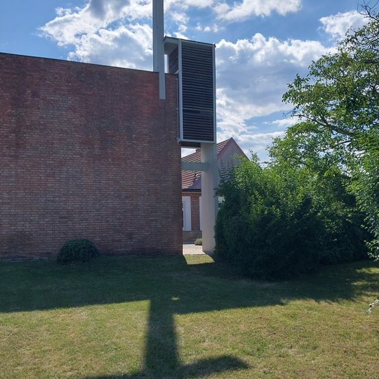 Ein Backsteingebäude mit Turm und Dachreiter wird von der Seite gezeigt, umgeben von Bäumen und Sträuchern. Der Himmel ist klar mit verstreuten Wolken.