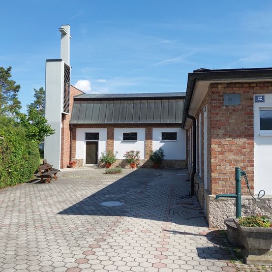 Ein Außenbereich mit einem Backsteingebäude mit grauem Dach und mehreren Fenstern. Links steht eine Bank und rechts eine Wasserpumpe. Vor dem Gebäude stehen Topfpflanzen.