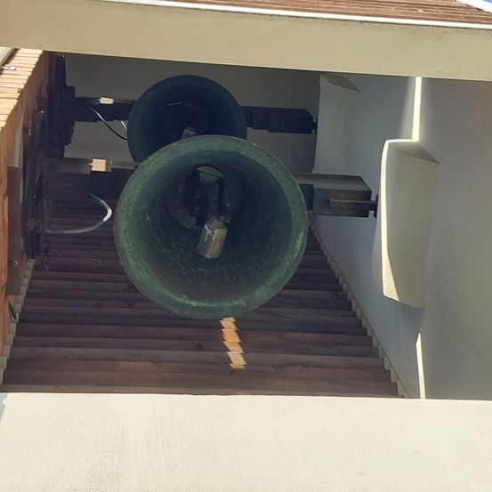 Eine große grüne Glocke ist auf einer Holzkonstruktion montiert, mit einem Seil und einem kleinen Metallstück im Inneren. Die Glocke befindet sich auf einer Treppe mit Holzstufen.