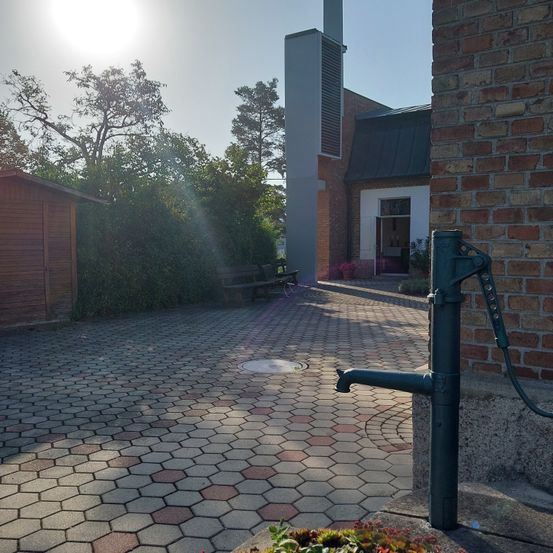 Ein Ziegelgebäude mit schwarzem Dach hat eine Pumpe und einen kleinen Garten. Bäume und Pflanzen umgeben das Gebiet. Eine Bank steht in der Nähe des Gebäudes.