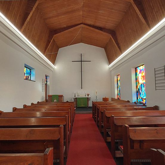 Der Innenraum einer Kirche zeigt hölzerne Kirchenbänke, eine Holzdecke, Buntglasfenster, ein Kreuz an der Wand und einen roten Teppich, der zum Altar führt.