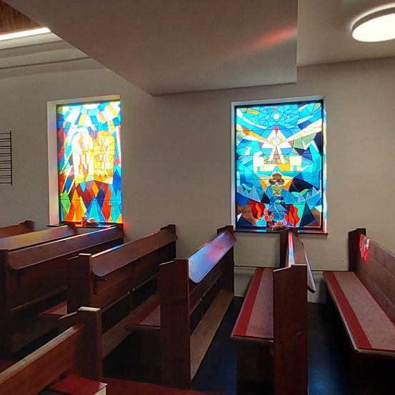 Ein Innenansicht einer Kapelle mit Buntglasfenstern und hölzernen Kirchenbänken. Die Fenster haben lebhafte Designs.
