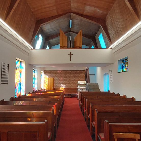 Eine leere Kirche mit Holzbänken, einem roten Teppich und Buntglasfenstern. Ein Kreuz ist an der Wand über den Bänken angebracht.