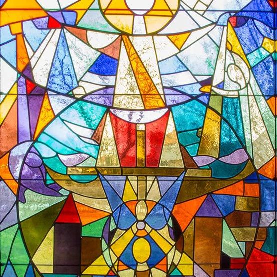 Ein Buntglasfenster mit einem Engel in der Mitte, dessen Flügel weit ausgebreitet sind, umgeben von leuchtenden geometrischen Formen in verschiedenen Farben.