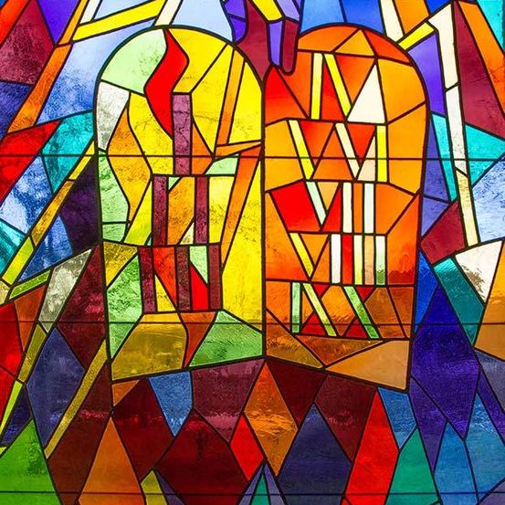 Ein farbenfrohes Buntglasfenster zeigt zwei Tafeln, möglicherweise die Zehn Gebote darstellend, umgeben von leuchtenden Farben und geometrischen Formen.