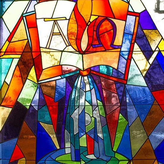 Ein Buntglasfenster zeigt ein offenes Buch mit einer Flamme darüber, umgeben von einem Mosaik aus verschiedenen farbigen Formen und Mustern.
