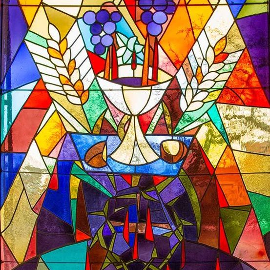 Ein Buntglasfenster zeigt einen Kelch mit Trauben, umgeben von Weizen, vor einem farbenfrohen geometrischen Hintergrund.