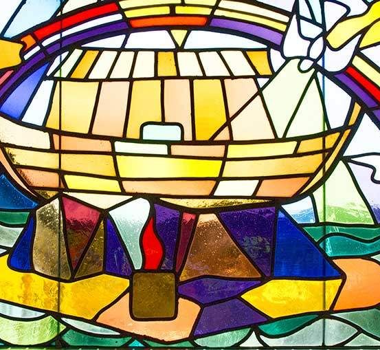 Ein Buntglasfenster zeigt ein Schiff, das auf einem Meer schwimmt, mit einem Regenbogen darüber. Das Schiff ist von farbenfrohen geometrischen Formen umgeben.