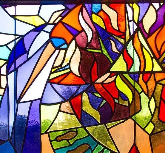 Ein farbenfrohes Buntglasfenster mit einem stilisierten Pelikan in Blau, hinter dem sich ein flammenartiges Muster in Rot und Gelb befindet, vor einem Hintergrund aus Blau- und Grüntönen.