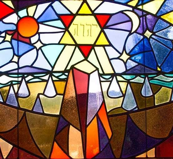 Ein Buntglasfenster zeigt einen leuchtenden Davidstern in der Mitte, umgeben von einem Regenbogen und himmlischen Elementen.
