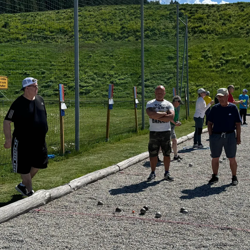 Eine Gruppe von Menschen steht in einem Außenbereich, möglicherweise spielen sie ein Spiel. Sie stehen auf einer Schotterfläche mit einigen verstreuten Bällen. Hinter ihnen befindet sich ein Grasfeld mit einem Zaun.