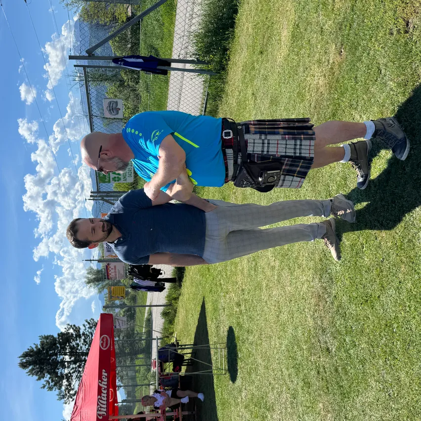 Zwei Männer in Freizeitkleidung stehen auf einem Grasfeld. Einer trägt ein Kilt und der andere eine Brille. In der Nähe befindet sich ein Zelt mit einem Schild, das 'Hacker' steht. Dahinter steht ein Zaun mit einem Banner. Der Himmel ist blau mit Wolken.