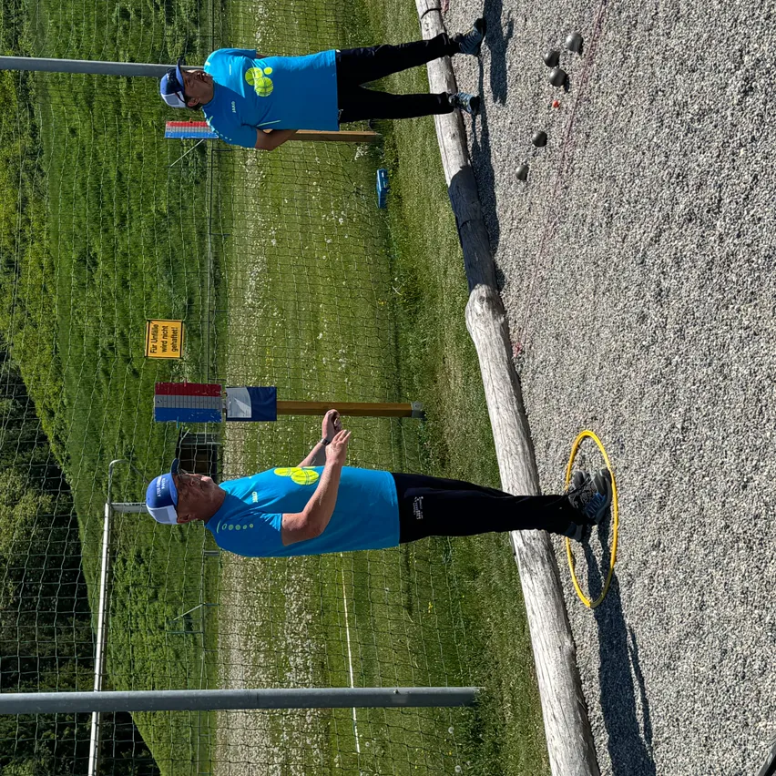 Zwei Männer spielen draußen auf einem Kiesplatz Boccia, einer liegt auf dem Boden und zielt, während der andere hinter ihm steht.