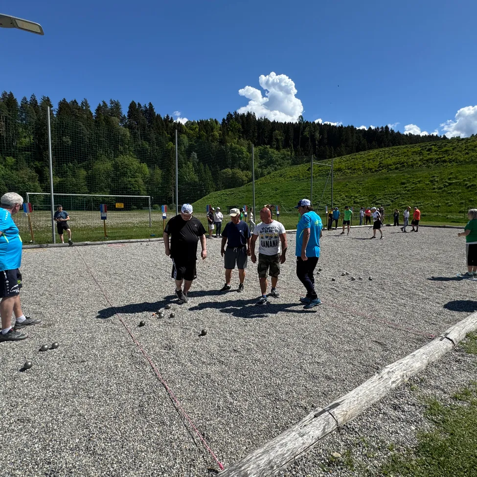 Eine Gruppe von Menschen spielt ein Spiel auf einem Schotterfeld, umgeben von einem grünen Hügel und einem blauen Himmel. Bäume und ein Fußballtor sind im Hintergrund zu sehen.