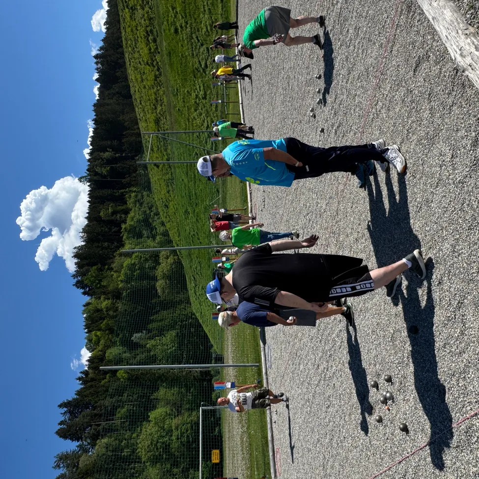 Eine Gruppe von Menschen spielt ein Spiel auf einem Kiesfeld mit einem grünen Feld und Bäumen im Hintergrund.