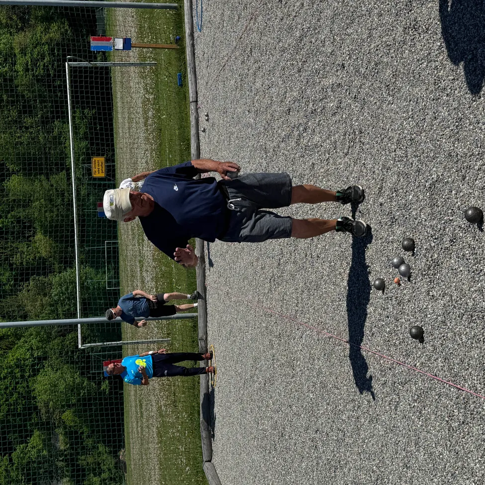Ein Mann bereitet sich auf den Wurf eines Balls in einem Spiel auf einem Kiesfeld vor. Andere stehen hinter ihm. Bäume und ein Netz sind im Hintergrund.
