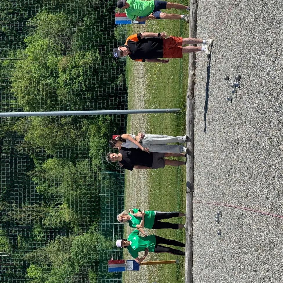 Fünf Personen in grünen und schwarzen Shirts spielen Boccia auf einem Kiesplatz mit Netz. Eine Person steht in der Nähe der Bälle.