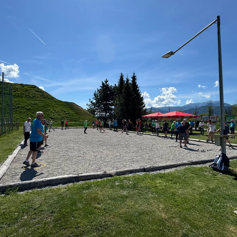 Eine Gruppe von Menschen spielt ein Spiel auf einem Kiesplatz. Sie tragen Turnschuhe und halten Stöcke. Im Hintergrund befinden sich Bäume und Berge.