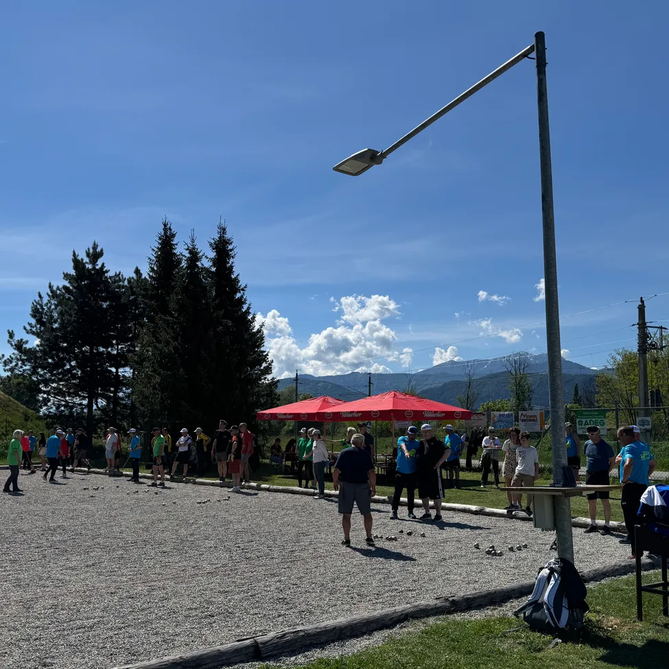 Eine Gruppe von Menschen versammelt sich im Freien in der Nähe eines roten Schirms und spielt Boule auf einem Kiesplatz. Bäume und Berge sind im Hintergrund unter einem blauen Himmel.