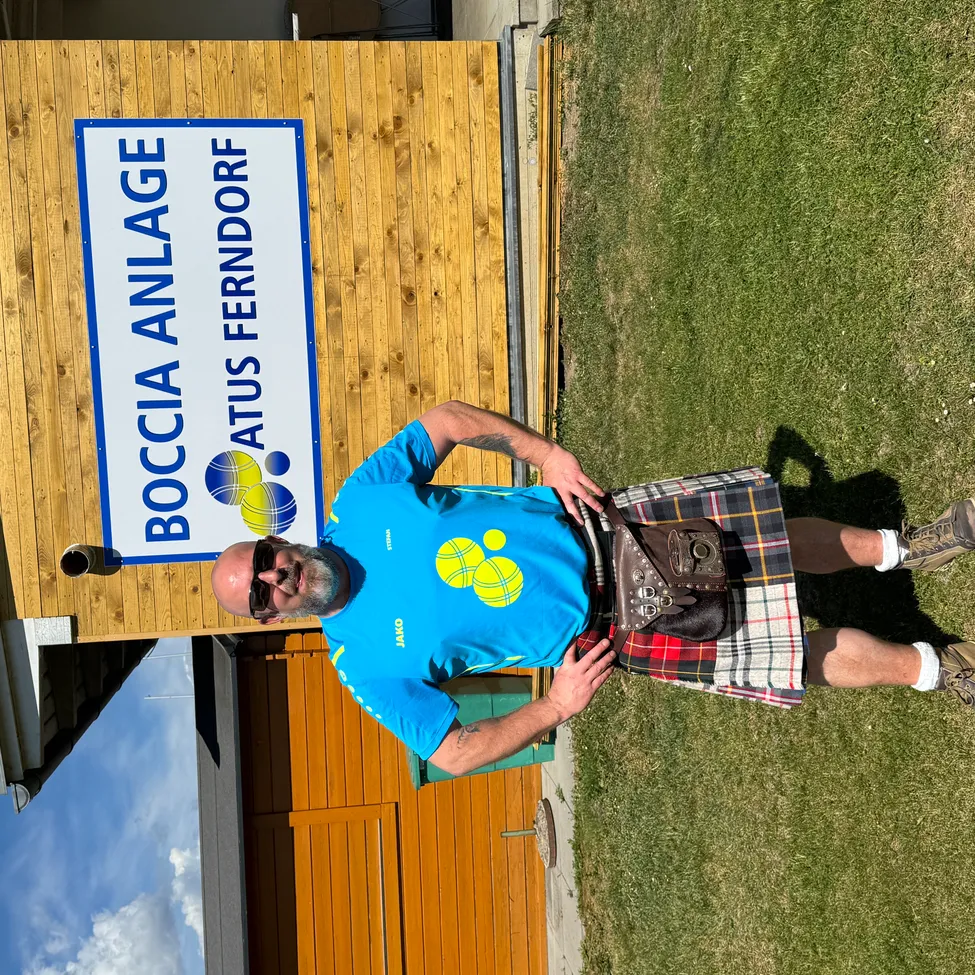 Ein Mann in einem blauen Shirt und einem Kilt posiert für ein Foto vor einem Boccia Anlage-Schild.