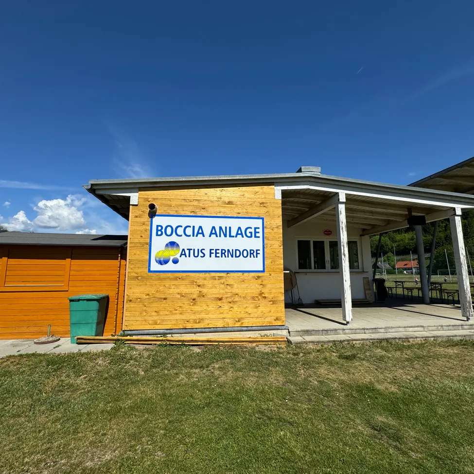 Ein kleines Gebäude mit einem Schild, das Boccia Anlage Atus Ferndorf steht. Es gibt einen grünen Mülleimer auf der linken Seite. Das Gebiet hat Gras und Bäume.