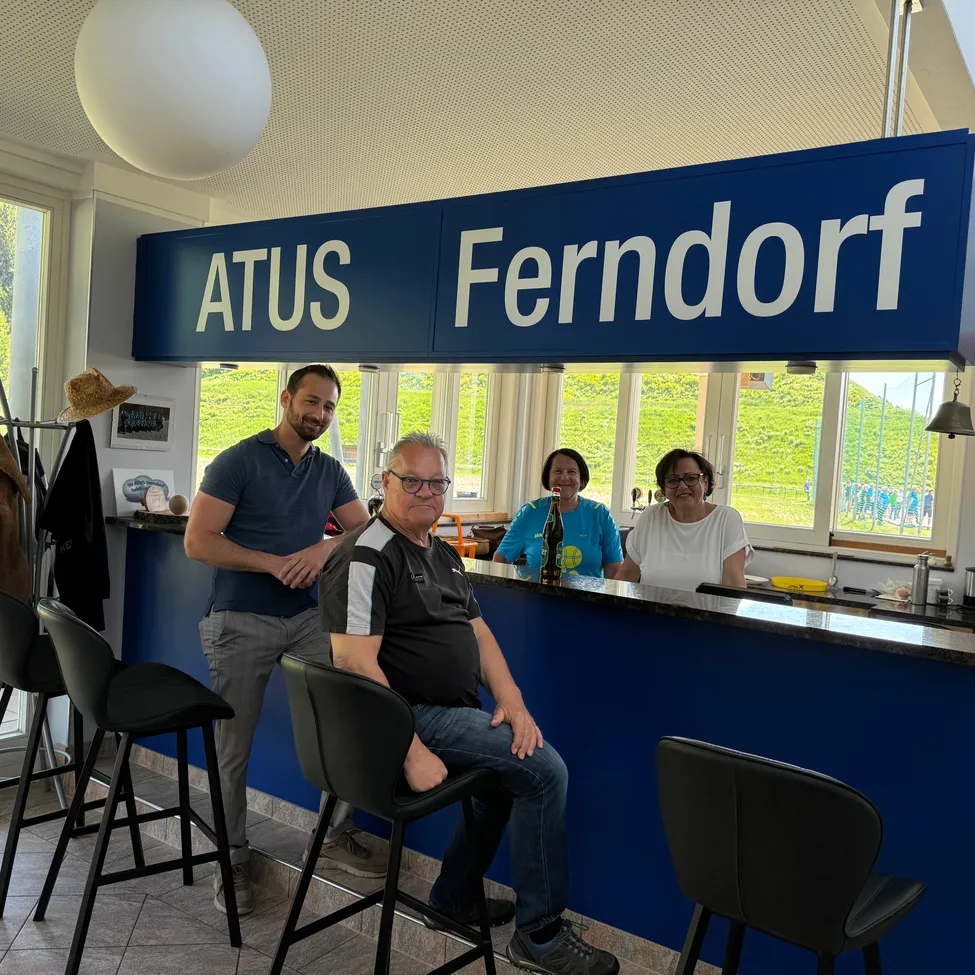 Drei Menschen sitzen an einer Bar und lächeln. Hinter ihnen steht ein blaues Schild, das ATUS Ferndorf besagt. Links steht ein Mann mit verschränkten Armen.