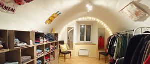 Ein kleiner, gewölbter Raum mit Holzboden und weißen Wänden. Der Raum hat einen Stuhl mit einem gelben Kissen, ein Regal mit gefalteter Kleidung, einen Spiegel und ein Fenster. Es gibt auch eine Heizung und dekorative Lichter.