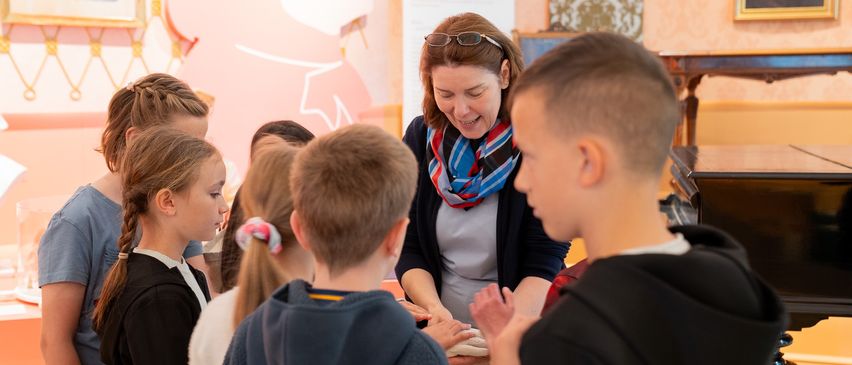 Eine Frau mit einer Brille auf dem Kopf unterrichtet eine Gruppe von Kindern in einem Handwerk, möglicherweise Brot herstellen.