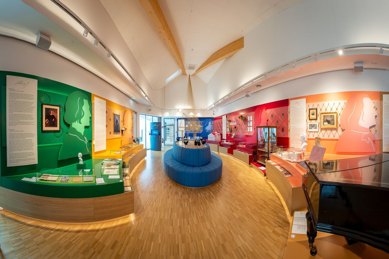 Eine moderne Museumsausstellung mit farbenfrohen Wänden, einem blauen runden Sitzbereich, Holzböden und verschiedenen Displays. Die Wände haben grüne, gelbe und rote Akzente mit Kunstwerken, Skulpturen und gerahmten Bildern.