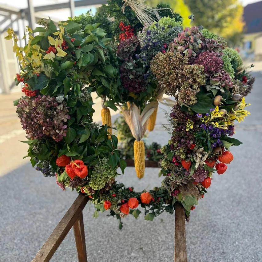 Ein Kranz aus verschiedenen Blumen, Blättern und Mais wird auf einem Holzständer im Freien ausgestellt.