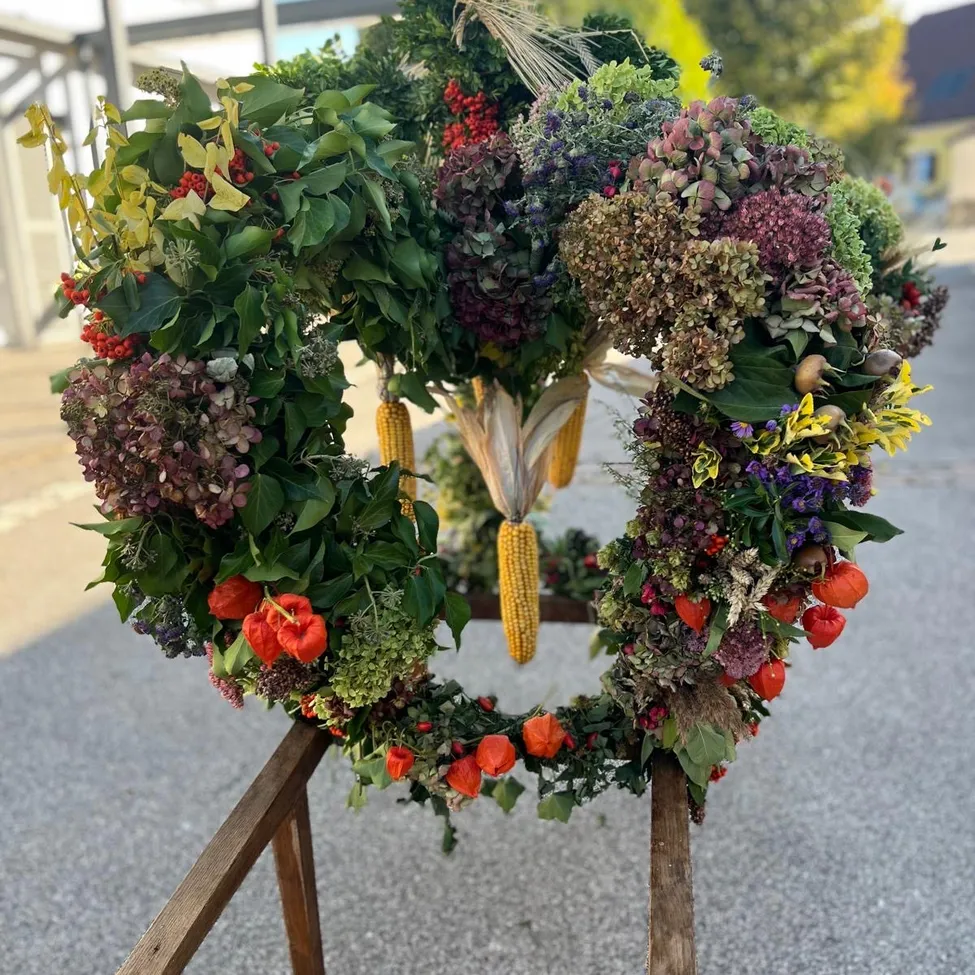Ein Kranz aus verschiedenen Blumen, Blättern und Mais wird auf einem Holzständer im Freien ausgestellt.