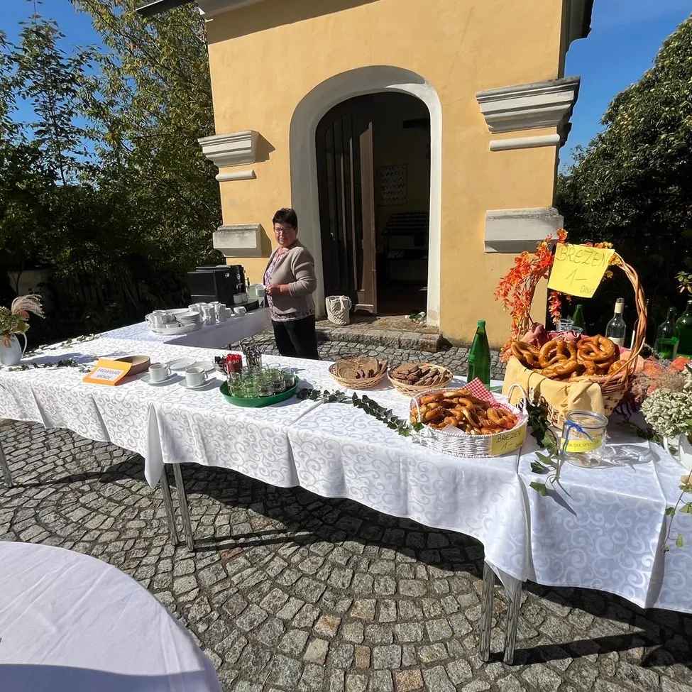 Eine Frau steht hinter einem Tisch voller Essen und Getränke, vor einem gelben Gebäude.