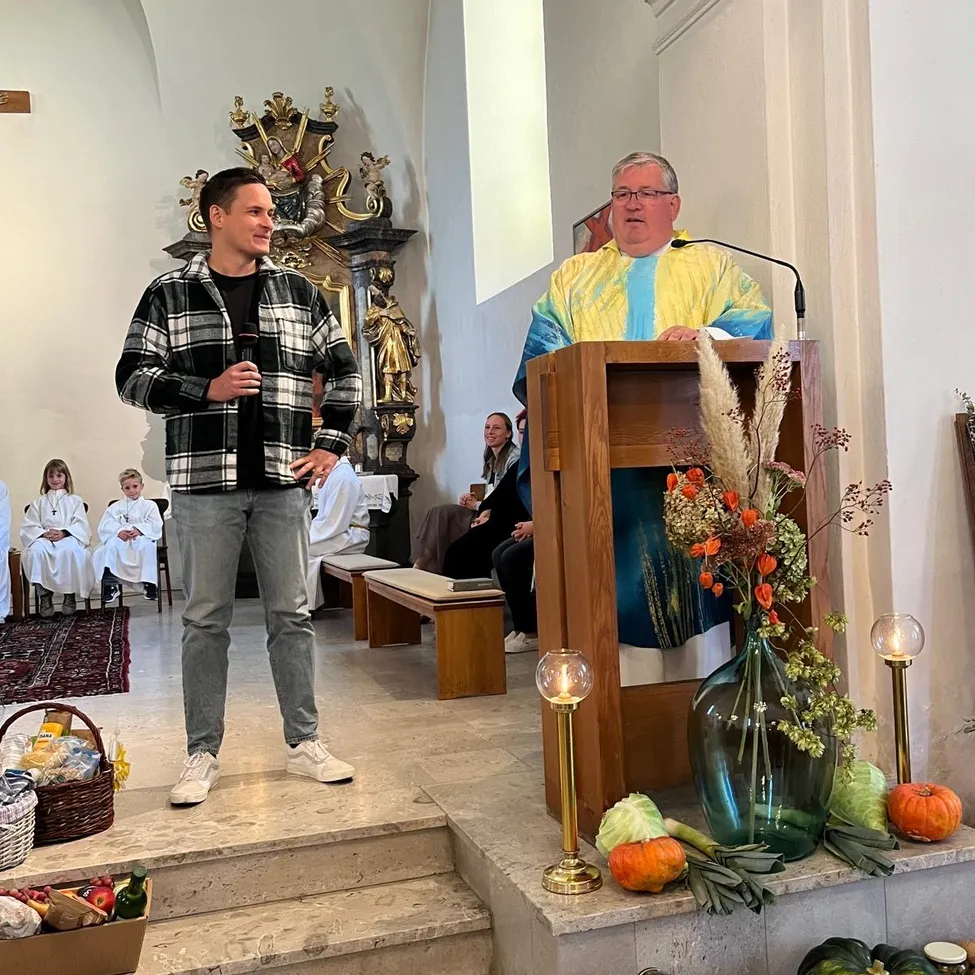 Ein Mann in einer karierten Jacke steht auf der Kirchenbühne, hält ein Mikrofon und spricht. Hinter ihm sitzt ein Priester in einem gelben Gewand an einem Rednerpult. Die Kirche ist mit Kürbissen, Blumen und Kerzen geschmückt.