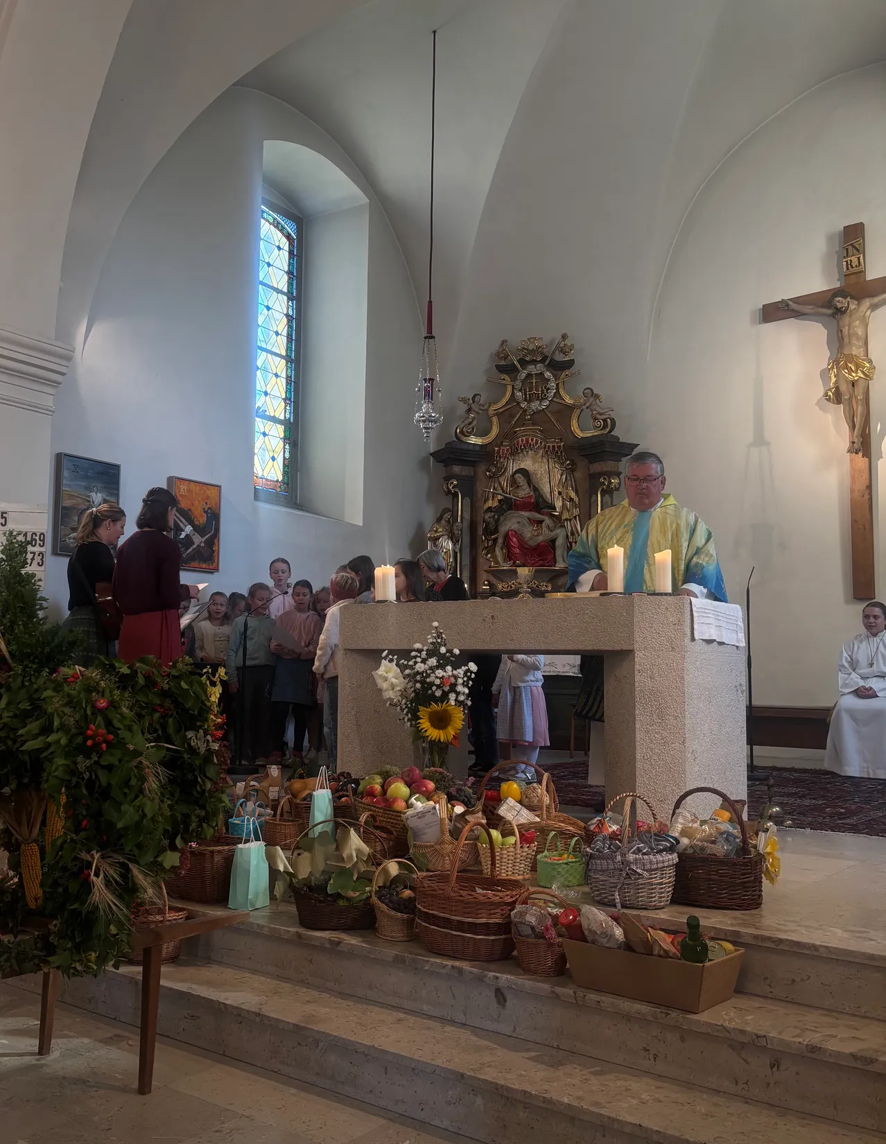 Ein Priester steht am Altar in einer Kirche, umgeben von Körben mit Früchten und Blumen, mit einem Kreuz an der Wand hinter ihm.